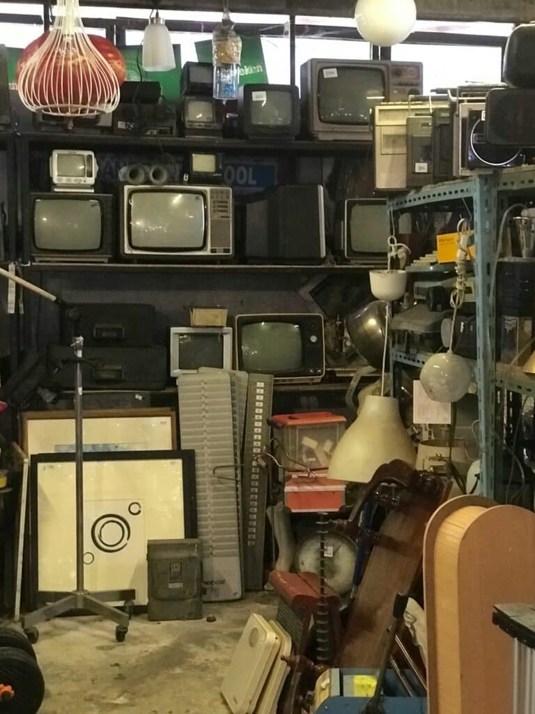 Téléviseurs CRT vintage : brocante d'électronique et objets. Collection de téléviseurs CRT vintage empilés sur des étagères métalliques, avec des boombox, éclairages et œuvres d'art dans un entrepôt.